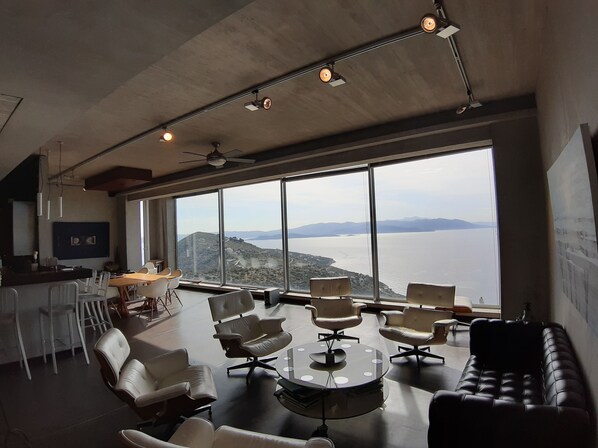 A modern interior with a panoramic view of a lake and mountains. It includes a dining area with white chairs and a round table, a sofa, and a bar area with stools. - ArtHouse (Marathon)