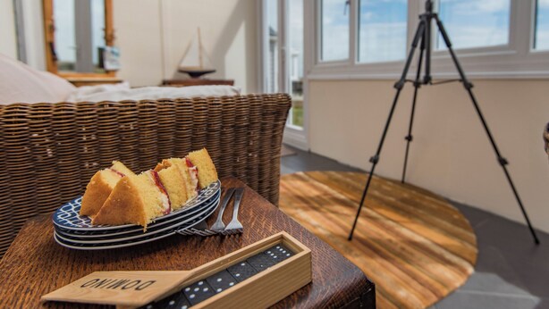 Une chaise en osier, une assiette remplie de parts de gâteau, un télescope sur un trépied et une boîte en bois avec une plaque nominative, le tout dans une pièce dotée de grandes fenêtres.