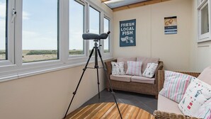 Une pièce dotée de grandes fenêtres, d’un télescope, d’un canapé en osier et d’une enseigne murale.