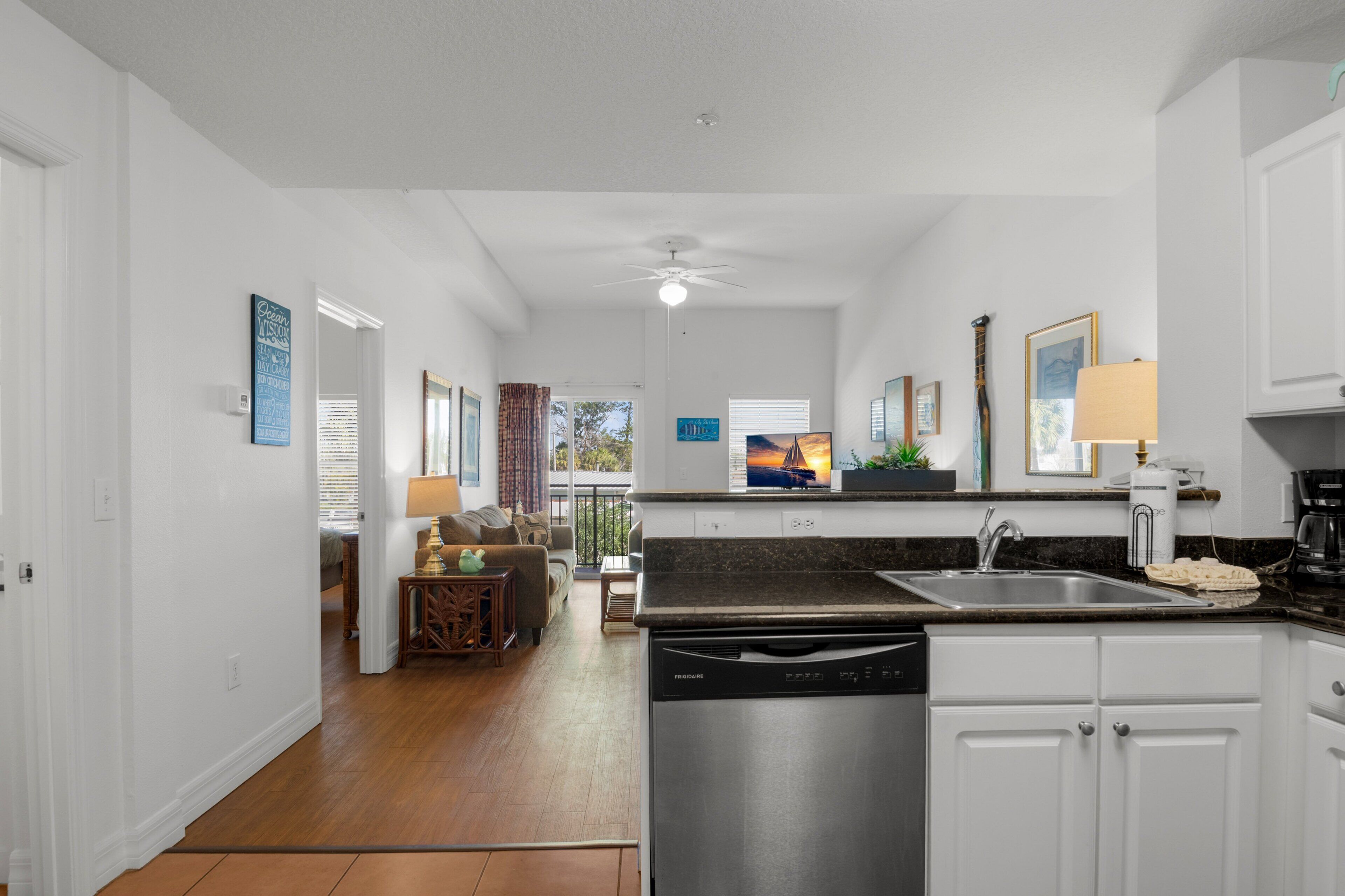 A modern kitchen with white cabinets, a dishwasher, and a sink. There is a dining area with a table and chairs, and a living room with a sofa and a coffee table.