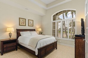 A bedroom with a large bed, two bedside tables with lamps, a wooden nightstand, a large window with shutters, and a view of the outdoors. - Modern 4 Bed 3 Bath Waterfront Home with Quick Gulf Access&Heated Pool (Cape Coral)