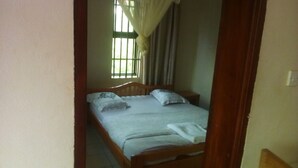 Una habitación pequeña con una cama individual, cabecera de madera, ventana con cortinas y mesita de noche.
