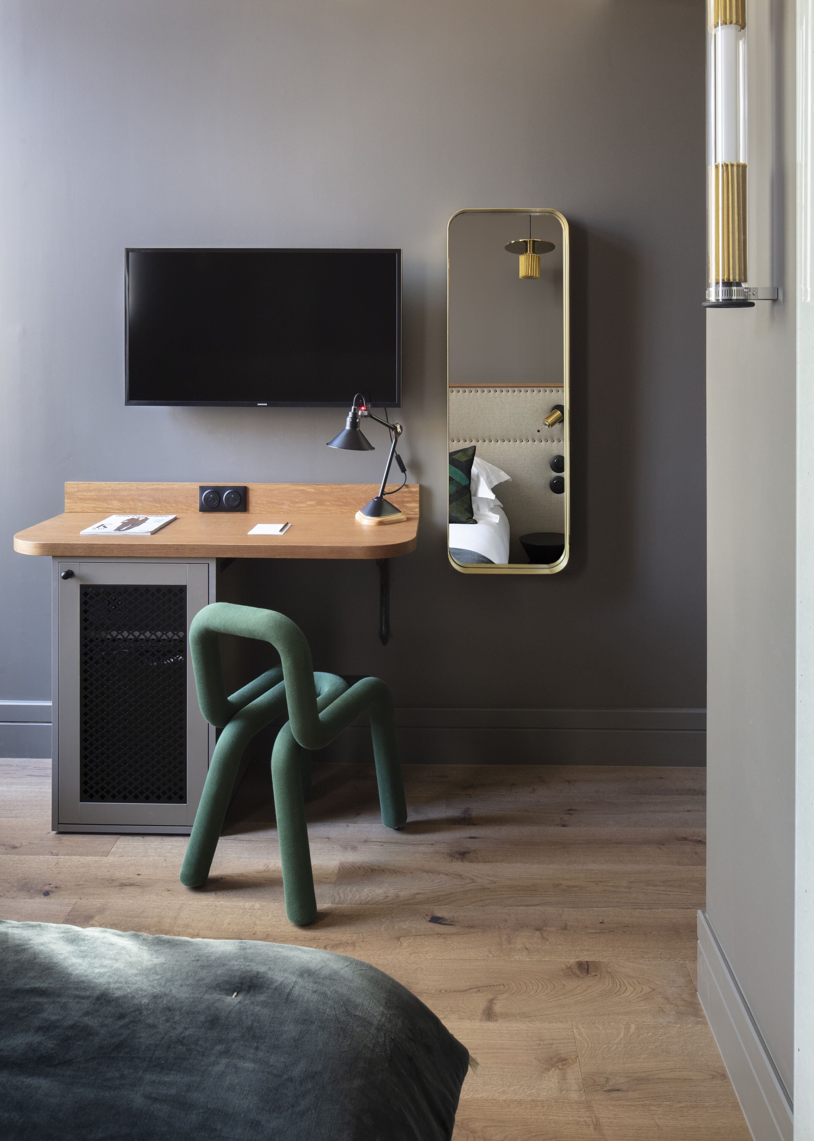 Une chambre avec un bureau en bois, une chaise verte, un lit, une télévision et un miroir.