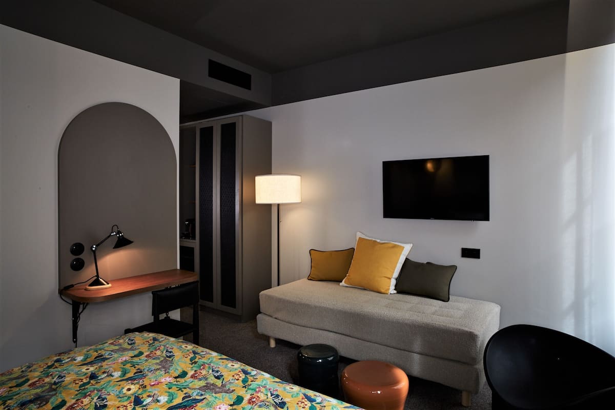 A hotel room with a bed, a desk, a chair, a sofa, a TV, and a lamp.
