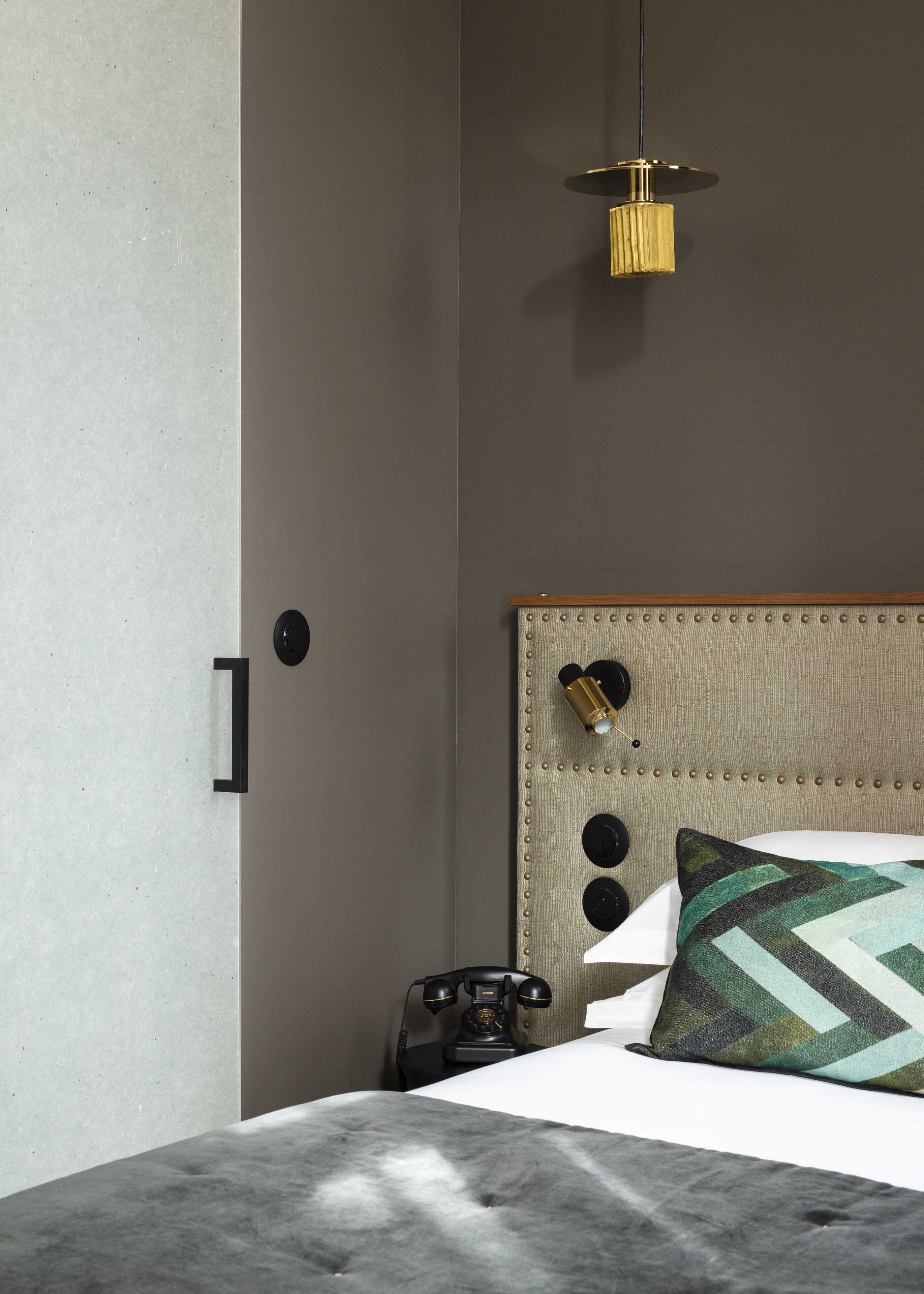 Un dormitorio moderno con cabecera gris, una lámpara colgante, un teléfono antiguo y un cojín estampado.