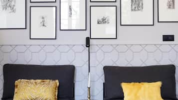 A room with two grey sofas, each with a yellow pillow, and a small table with a candle. The wall above the sofas is decorated with framed black and white sketches.