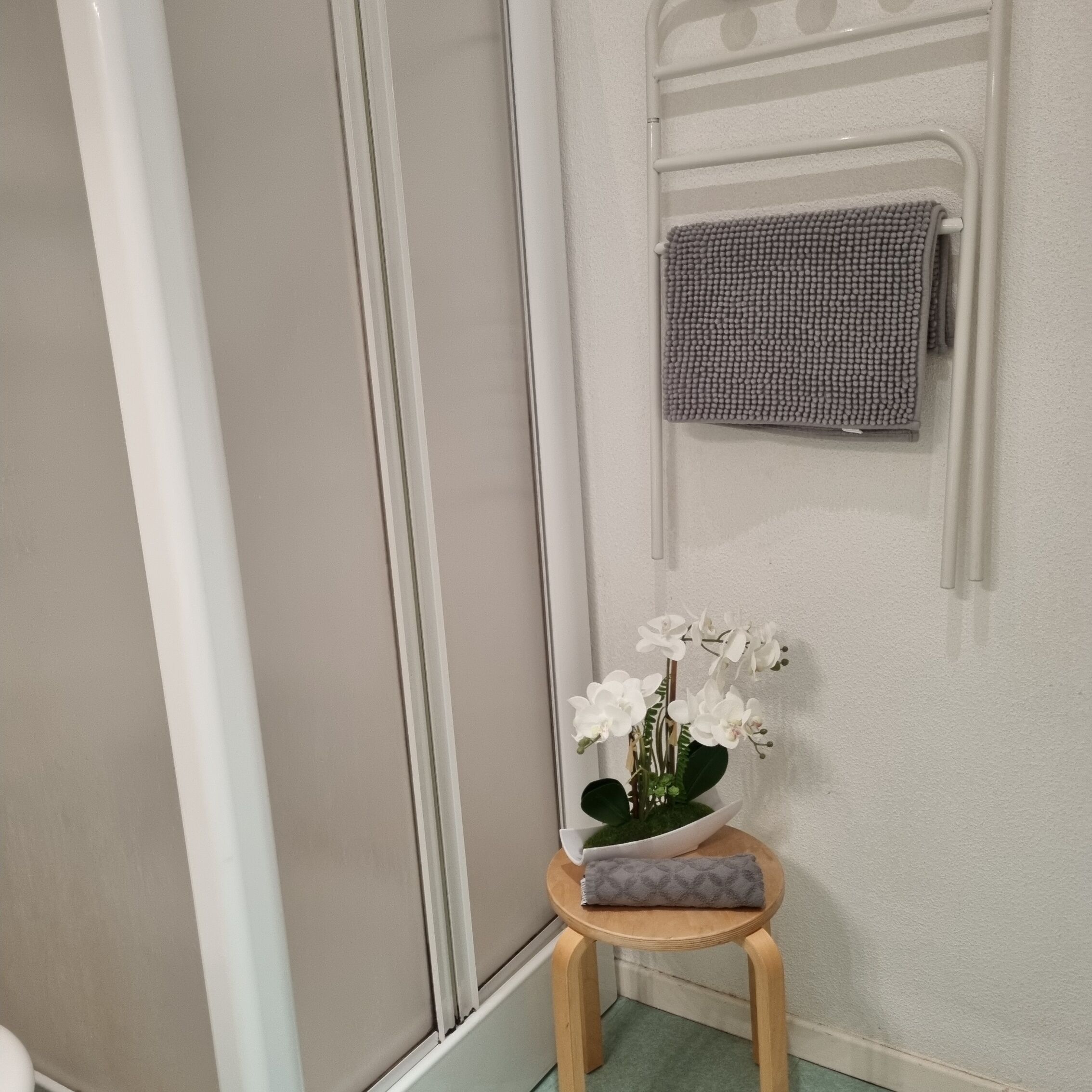Een badkamer met een witte douche, een handdoekrek met een grijze handdoek en een klein krukje met een potorkidee.