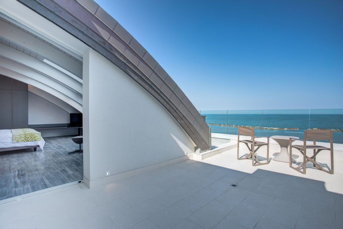 A modern balcony with a bed, two chairs, and a view of the sea.