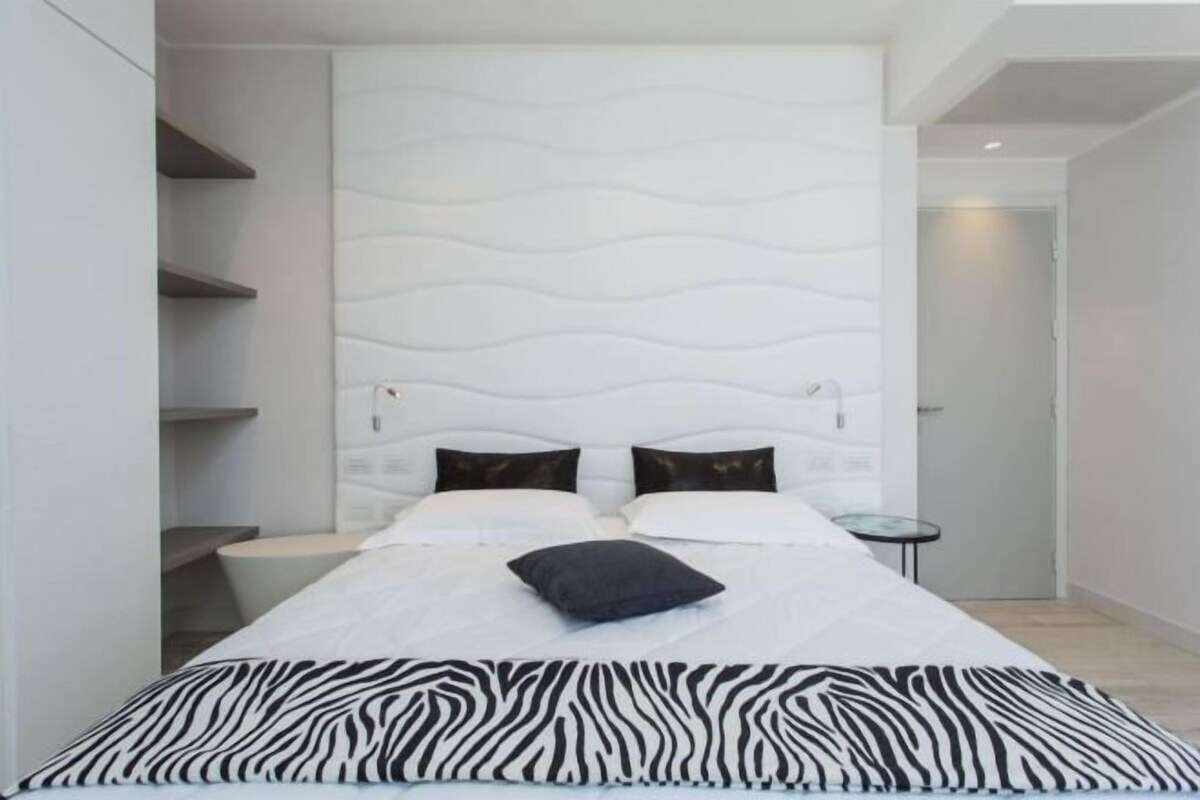 A modern bedroom with a white headboard, two black pillows, a zebra-patterned bedspread, and a small round table beside the bed.