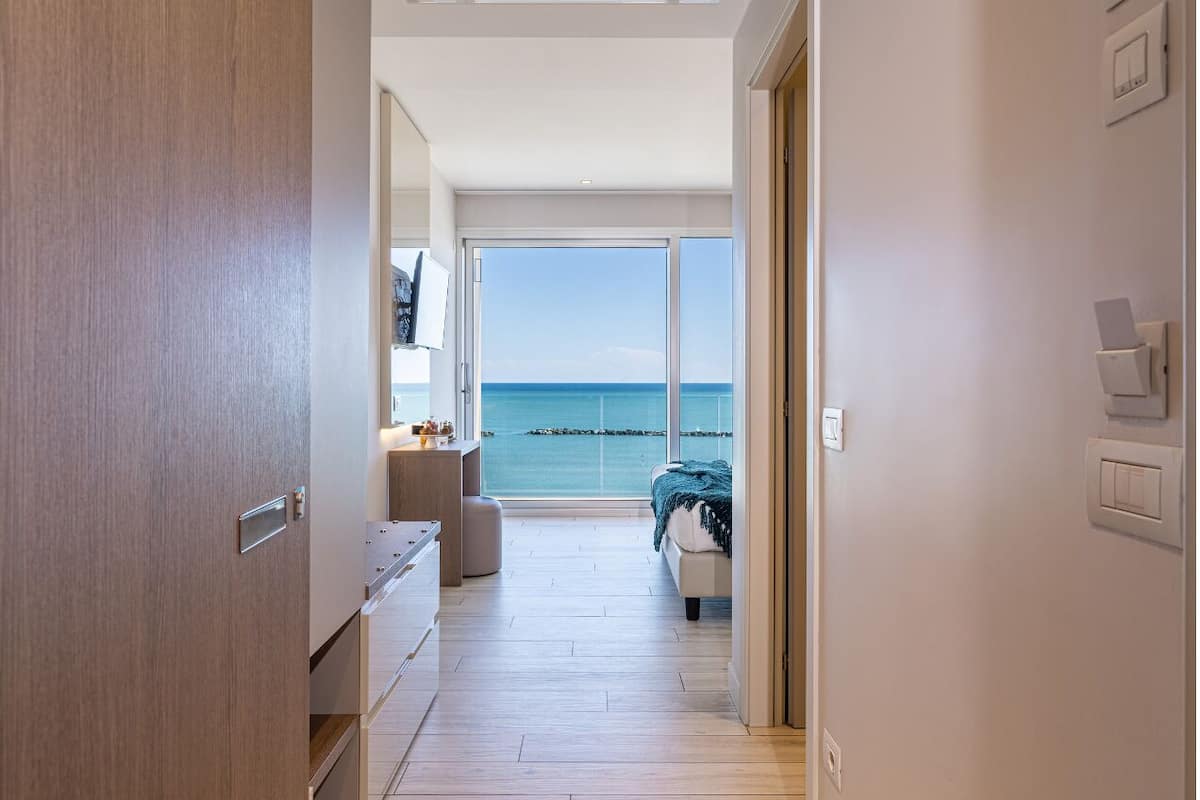 Een moderne hotelkamer met een houten vloer, een bed met een blauwe deken, een bureau en uitzicht op zee door grote ramen.