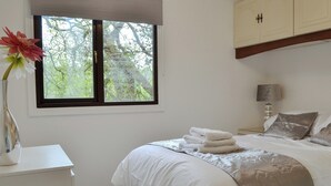 A bedroom with a bed, a nightstand, a lamp, a window with a view of trees, and a cabinet. - 2 bedroom accommodation in Benllech (Benllech)