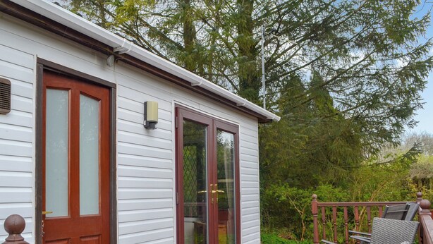 A white cabin with a wooden deck, a glass door, and a small outdoor seating area with wicker chairs. - 2 bedroom accommodation in Benllech (Benllech)