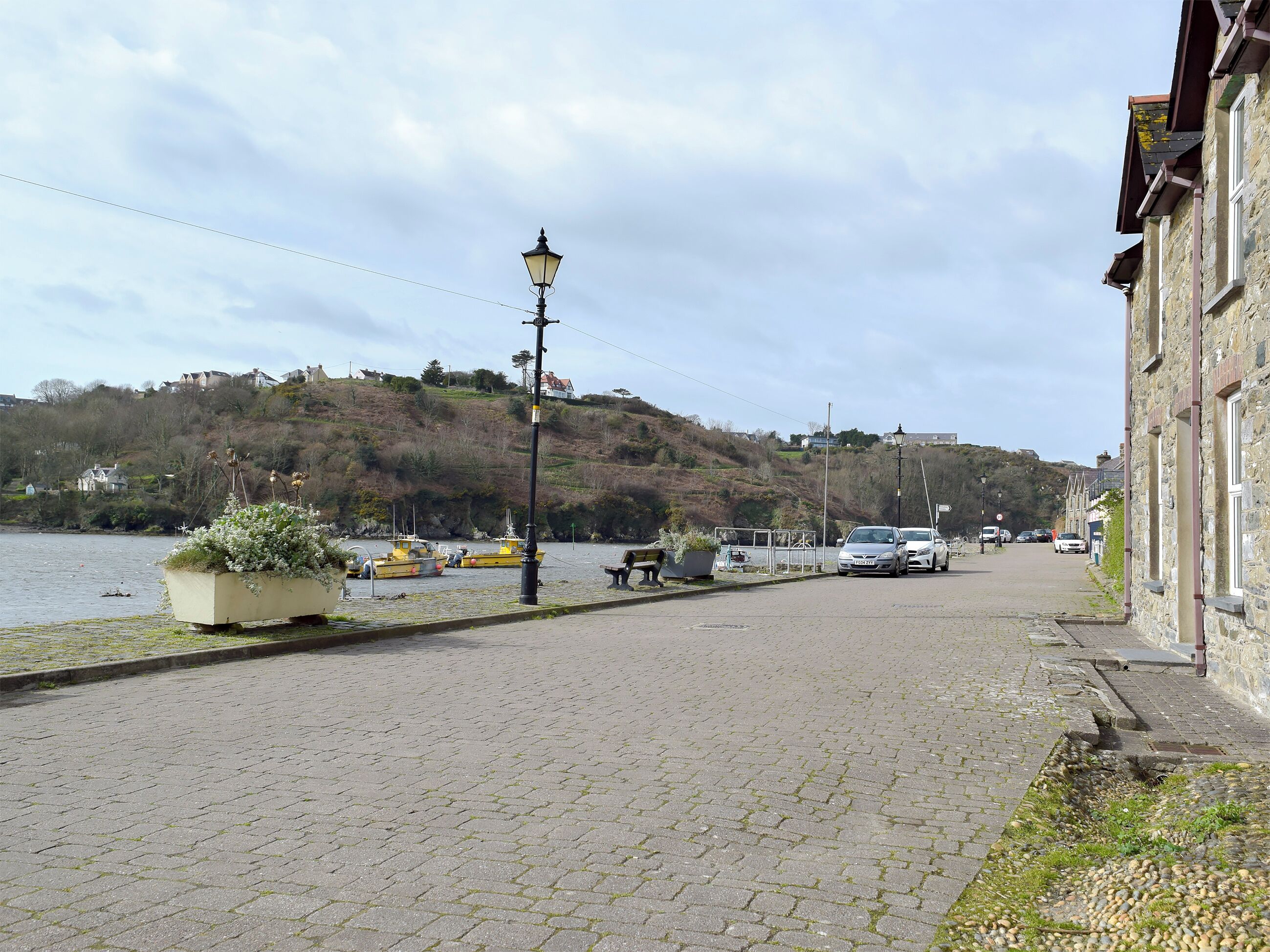 A cobblestone waterfront street with boats docked, a lamp post, and stone buildings.
