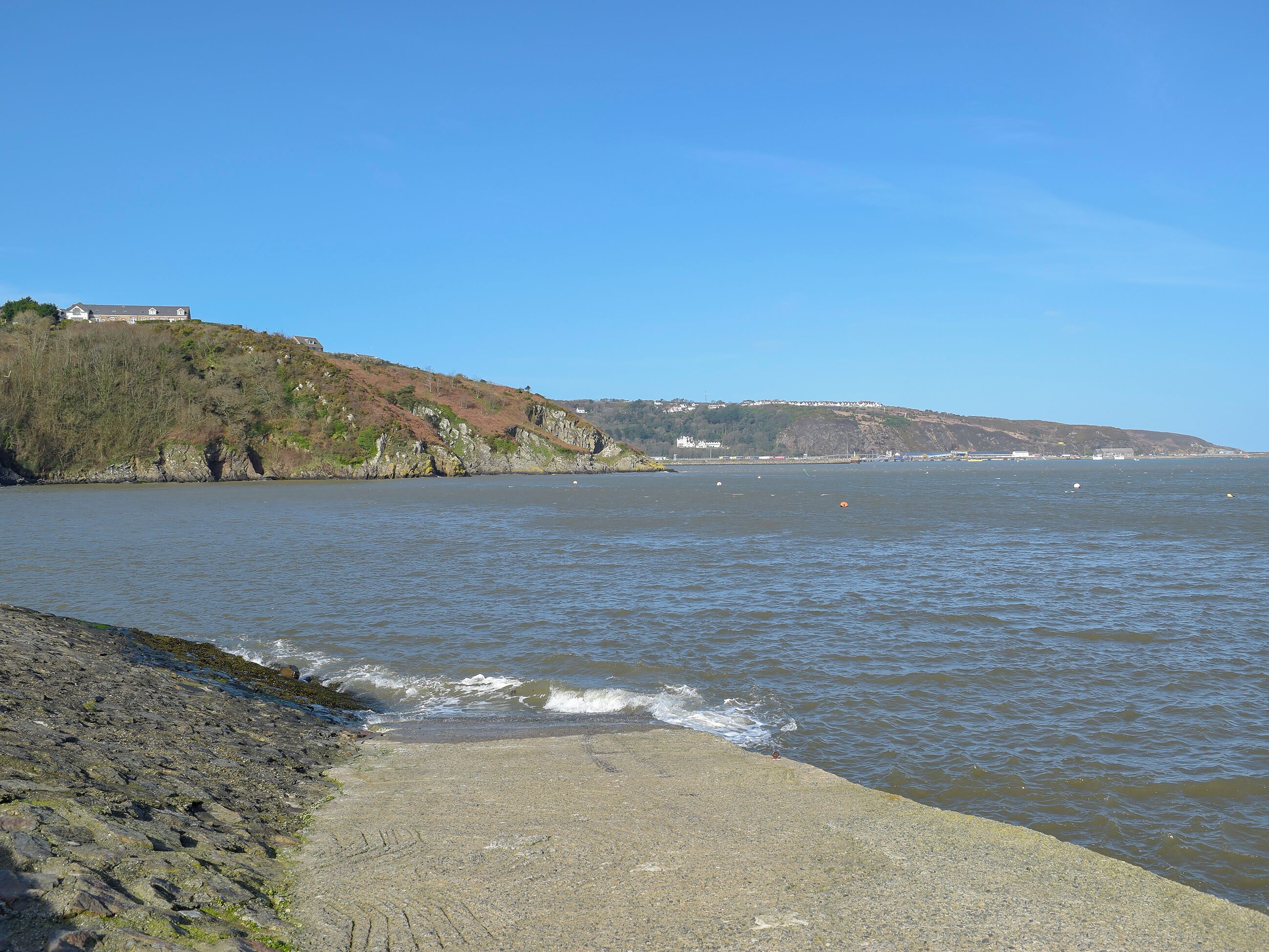 A coastal view with a rocky shoreline, calm water, and a distant hill with buildings.