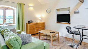 Een woonkamer met een bank, een houten salontafel, een aan de muur bevestigde televisie, en een eethoek met een stoel en tafel.