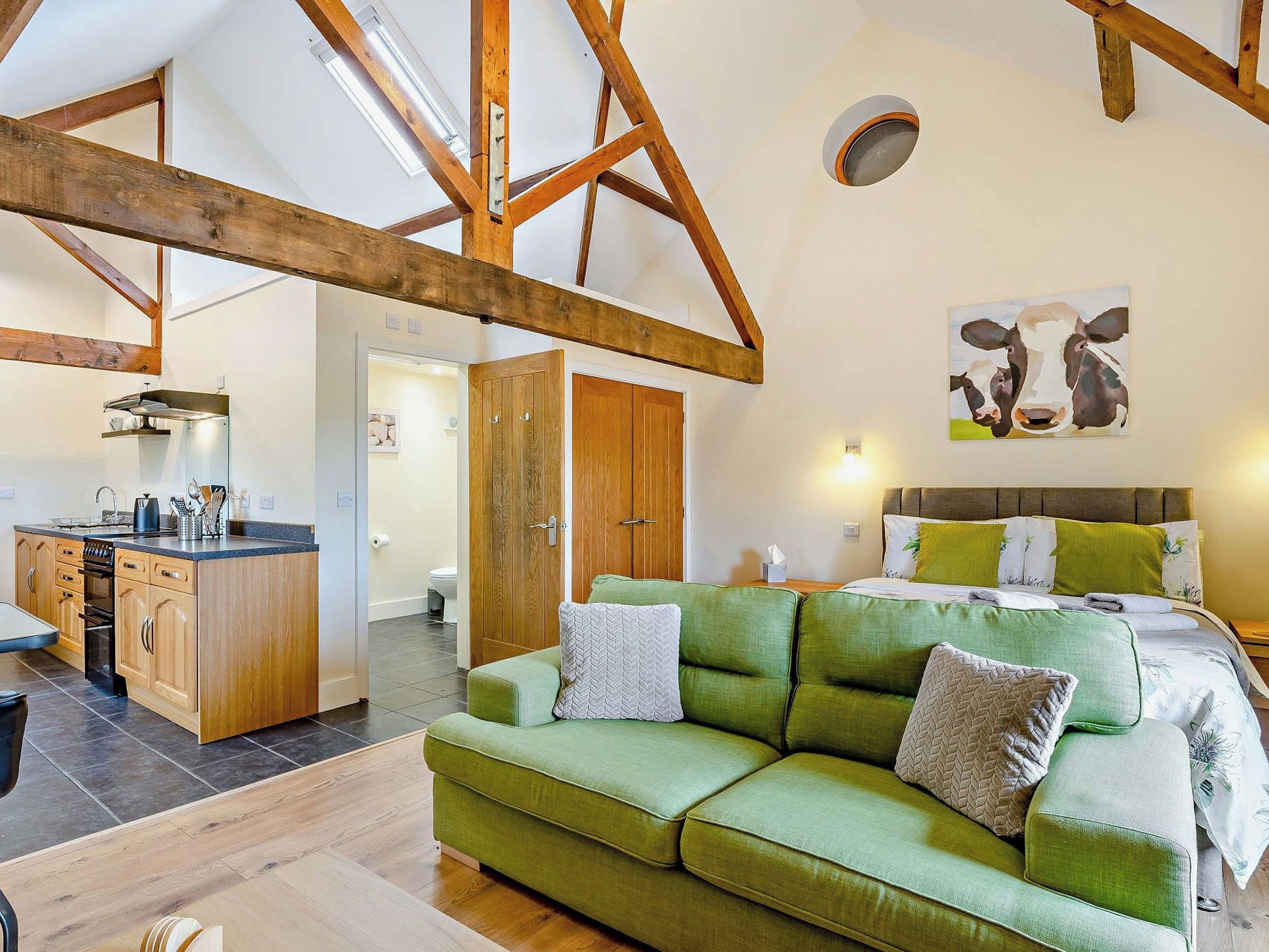 A cozy room with a green sofa, a bed with green and white bedding, a wooden ceiling, and a painting of cows on the wall.