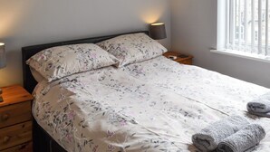 Ein ordentlich bezogenes Bett mit Blumenmuster, eine Nachttischlampe und ein Fenster mit Jalousien.