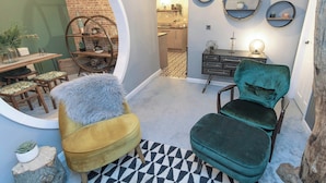 A cozy living room with a round mirror, a yellow armchair, a green velvet chair, and a dining area with a brick wall and wooden furniture.