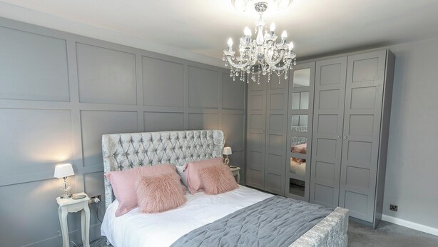 A bedroom with a tufted headboard, a grey bedspread, fluffy pillows, a chandelier, and a grey wardrobe with mirrored doors.