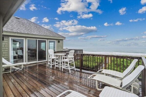 A wooden deck with a glass door leading to a room, outdoor seating, and a view of the ocean.