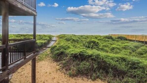 A wooden pathway leading through a sandy area with dense green vegetation, under a clear sky. - Fabulous Find - Spendlove Oceanfront (Duck)