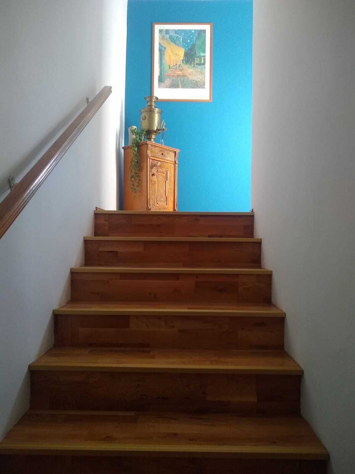 A wooden staircase with a handrail on the left, leading to a blue wall with a framed painting.