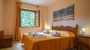 A hotel room with a bed, a nightstand, a desk, a window with curtains, and a painting on the wall. - Anticheterme / Trilocale (Sorano GR)