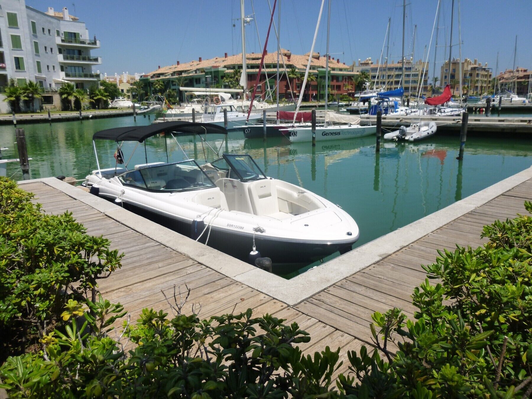 Un port de plaisance où un bateau à moteur blanc est amarré à un quai en bois, entouré de verdure et avec des bâtiments en arrière-plan.