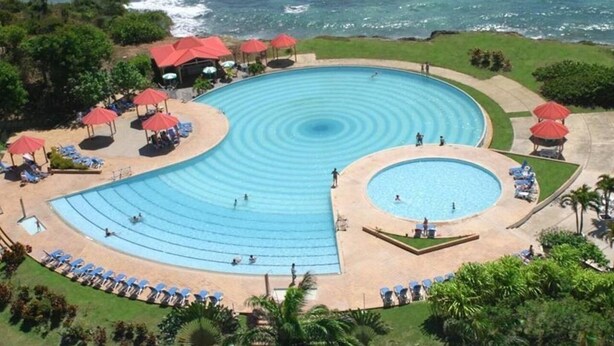 A large, circular swimming pool with multiple smaller pools, surrounded by lounge chairs and umbrellas, with a view of the ocean. - Nice furnished studio with small sea view<br><br><br> (SAINT FRANCOIS)