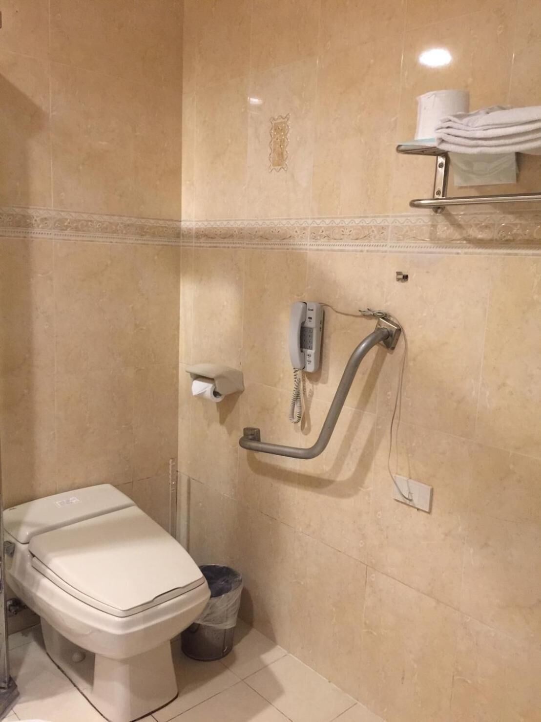 A bathroom with a toilet, a handrail, a telephone, and a trash bin.
