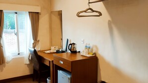 A hotel room with a wooden desk, a small refrigerator, a hanging coat rack, and a window with curtains.