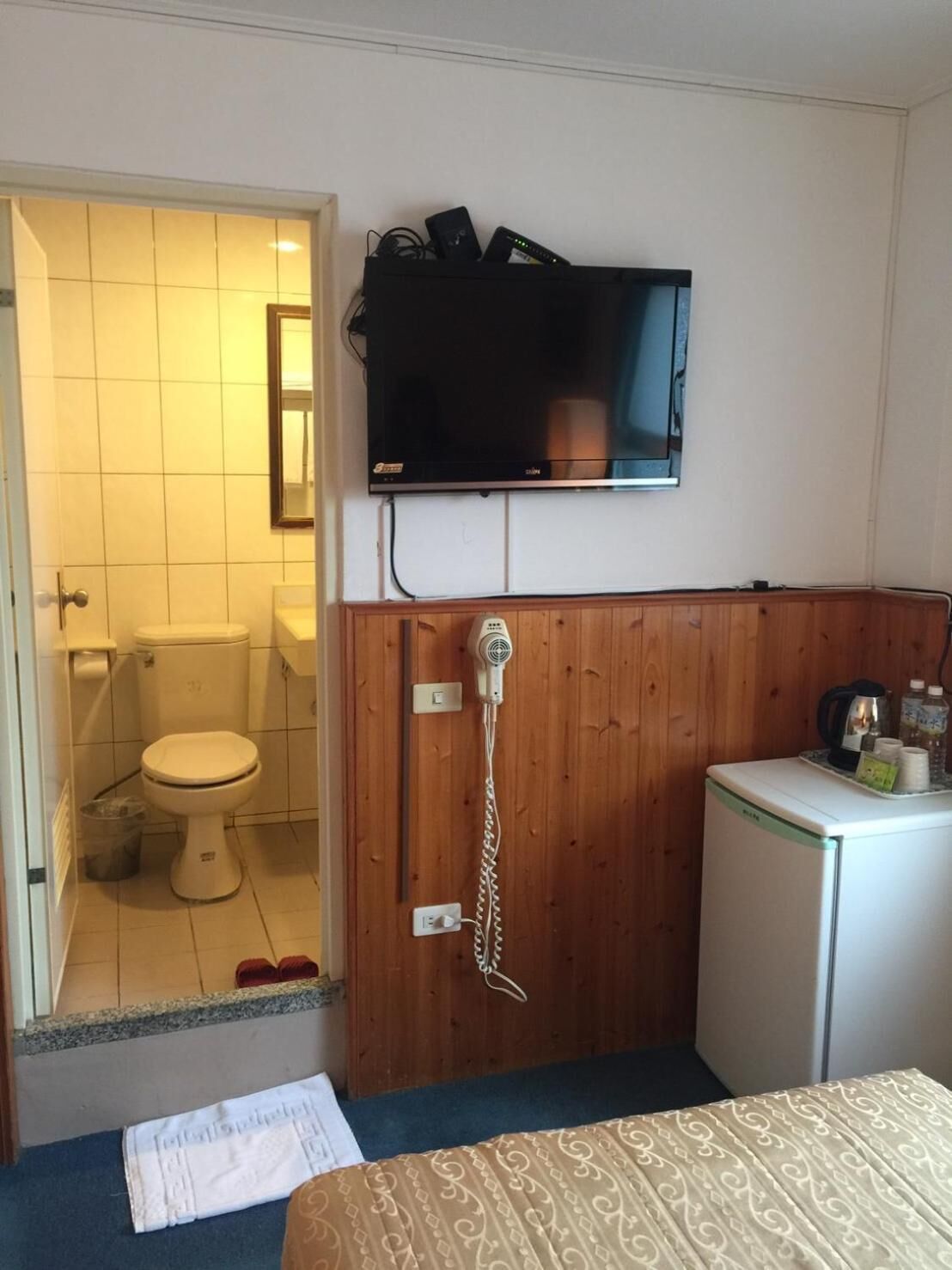 A small hotel room with a wooden panel wall, a television mounted above a wooden cabinet, a white toilet, and a small refrigerator.