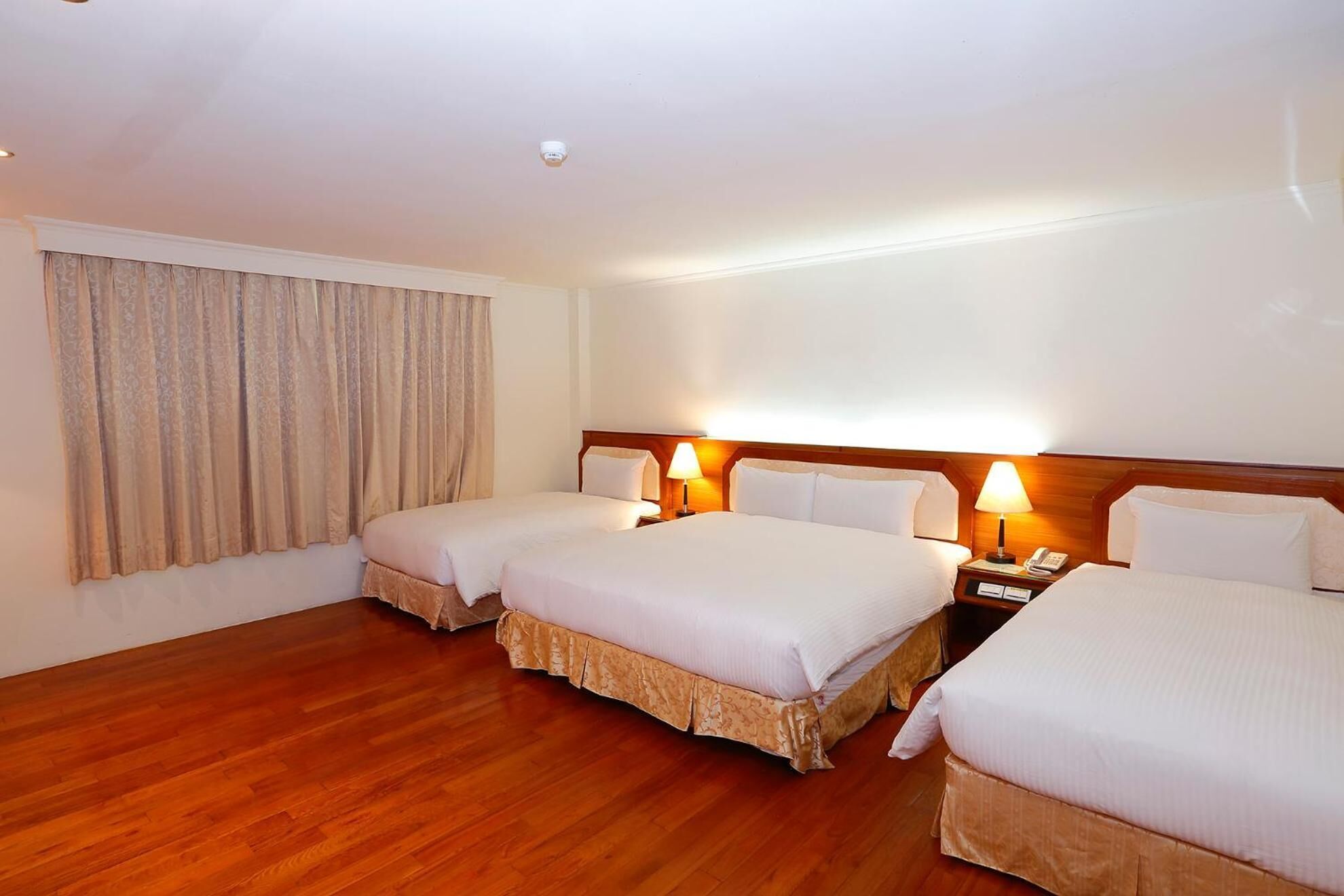 A hotel room with three single beds, wooden flooring, a large window with curtains, and bedside lamps.