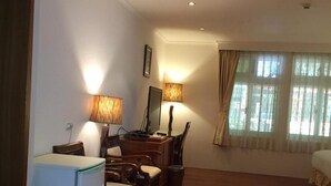 A cozy room with a wooden floor, a desk with a chair, a small refrigerator, and a bed with a window.