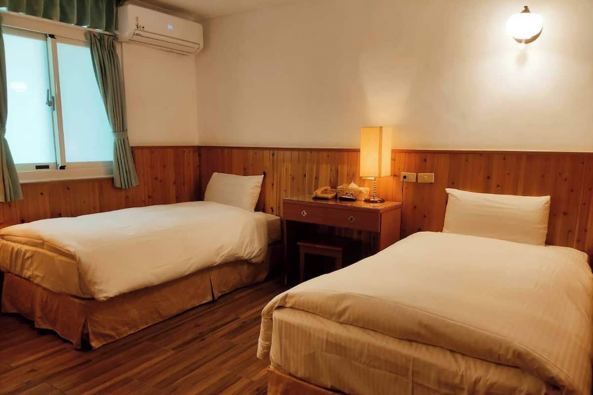 Habitación de hotel con dos camas individuales, suelo de madera, un escritorio con lámpara y una ventana con cortinas.
