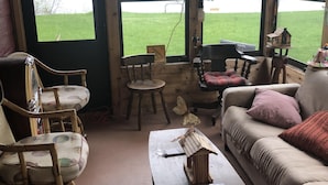 A cozy room with a wooden interior, a sofa, a rocking chair, a table with a model house, and large windows overlooking a grassy area. - NEWER FAMILY 3 BEDROOM 2 BATH LAKE CABIN (Swan River)