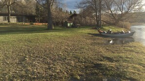 A grassy area with a boat docked by a river, a small shed, and a house in the background. - Best Location on the MIssouri River (Cascade)