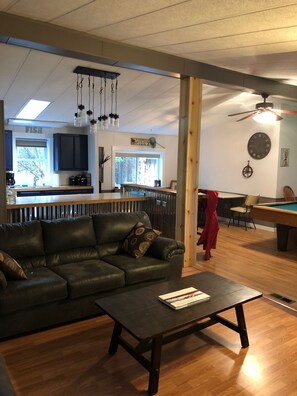 Een woonkamer met een donker leren bank, een houten salontafel, een pooltafel en een keuken op de achtergrond.