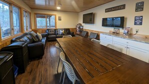 Interior acogedor de una cabaña con una mesa de comedor de madera, un sofá de cuero y una zona de cocina con televisión y cafetera.