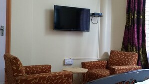 Quarto de hotel com televisão de tela plana na parede, duas poltronas estampadas, uma mesinha de madeira e uma tomada.