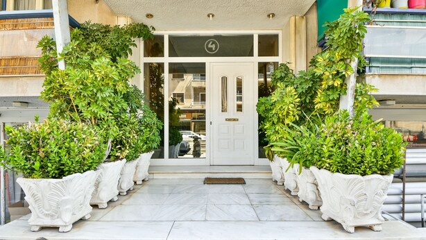 Entrada de hotel con puerta blanca, un pequeño balcón y plantas en macetas a ambos lados.
