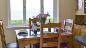 Un coin repas avec une table dressée pour deux personnes, un vase rempli de fleurs et une vue sur la mer grâce à de grandes fenêtres.