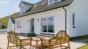 Une maison à deux étages, avec une façade blanche, un toit en tuiles et un porche à l’avant, agrémenté de deux bancs en bois.