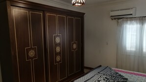 Ein Schlafzimmer mit großem Kleiderschrank, einem Bett mit gemusterten Betttextilien, einem Fenster mit durchsichtigen Vorhängen und einer Klimaanlage.