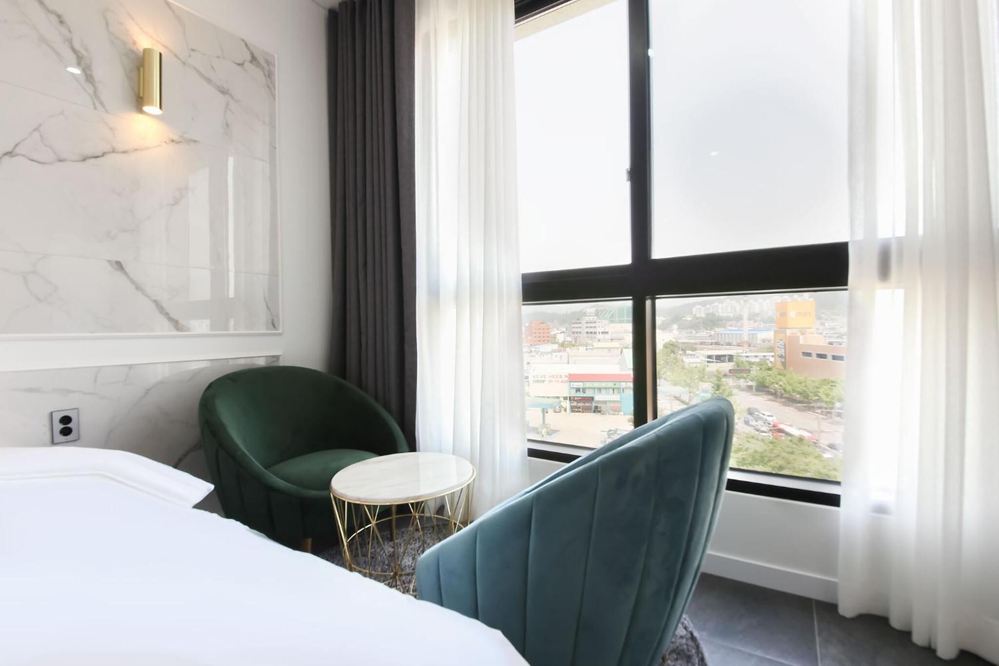 A hotel room with a bed, a green chair, a small round table, and a large window with a city view.