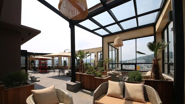 A rooftop terrace with wicker furniture, potted plants, and a glass ceiling offering a city view. - Hotel RAUM (Suncheon)