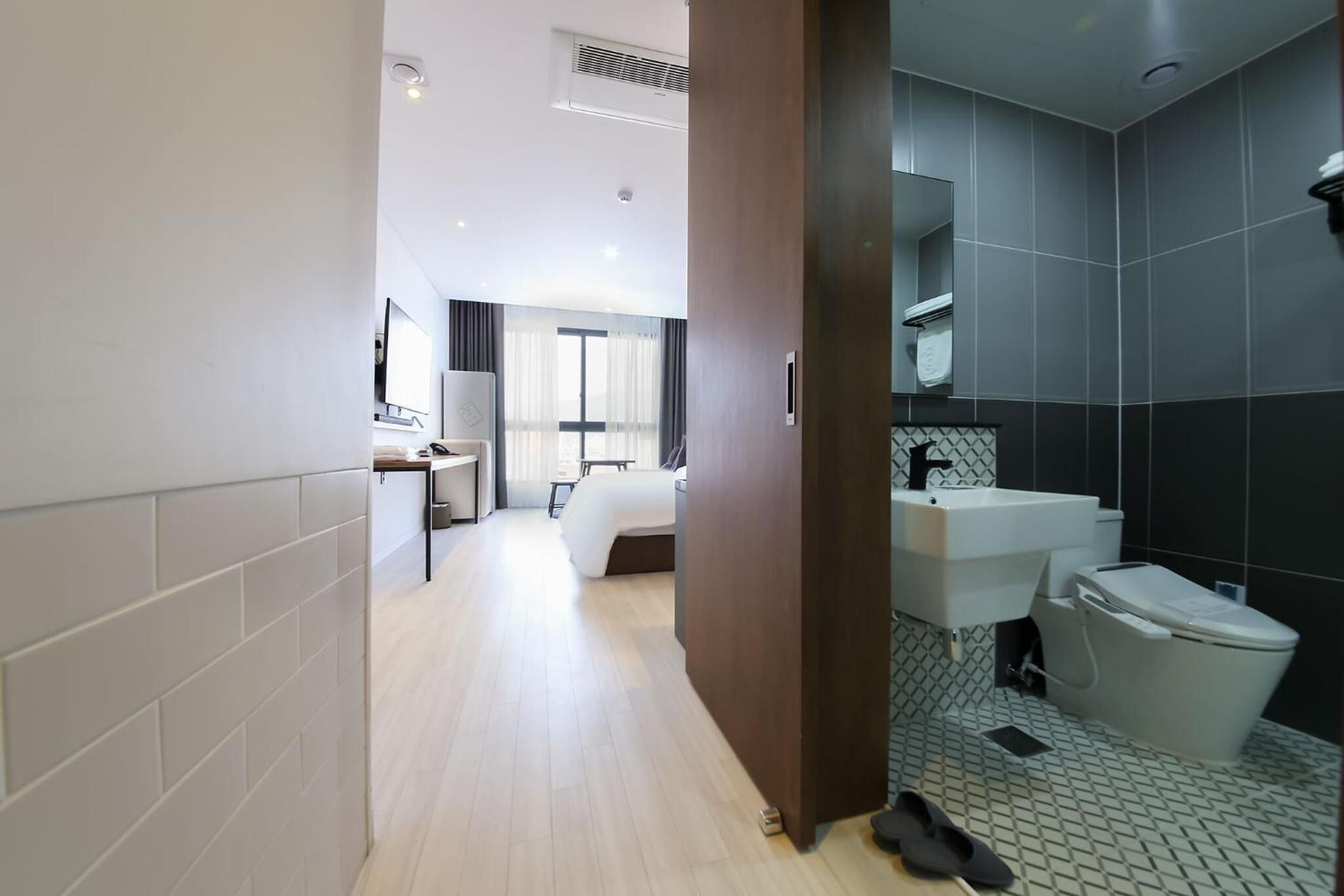 A modern hotel room with a wooden floor, a bed, a desk with a chair, a large window with curtains, a bathroom with a sink, mirror, and toilet.