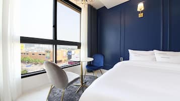 Habitación de hotel con una cama grande, un escritorio, una silla y vistas de la ciudad a través de un ventanal.