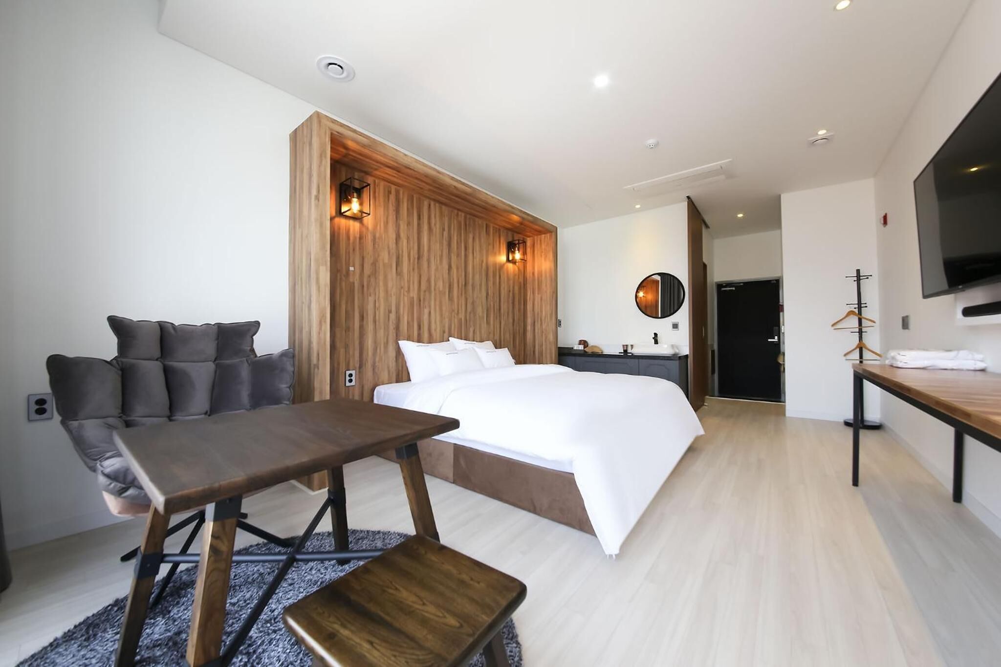 A modern hotel room with a large bed, a wooden accent wall, a seating area with a table and chairs, a flat-screen TV mounted on the wall, and a mirror above the bed.