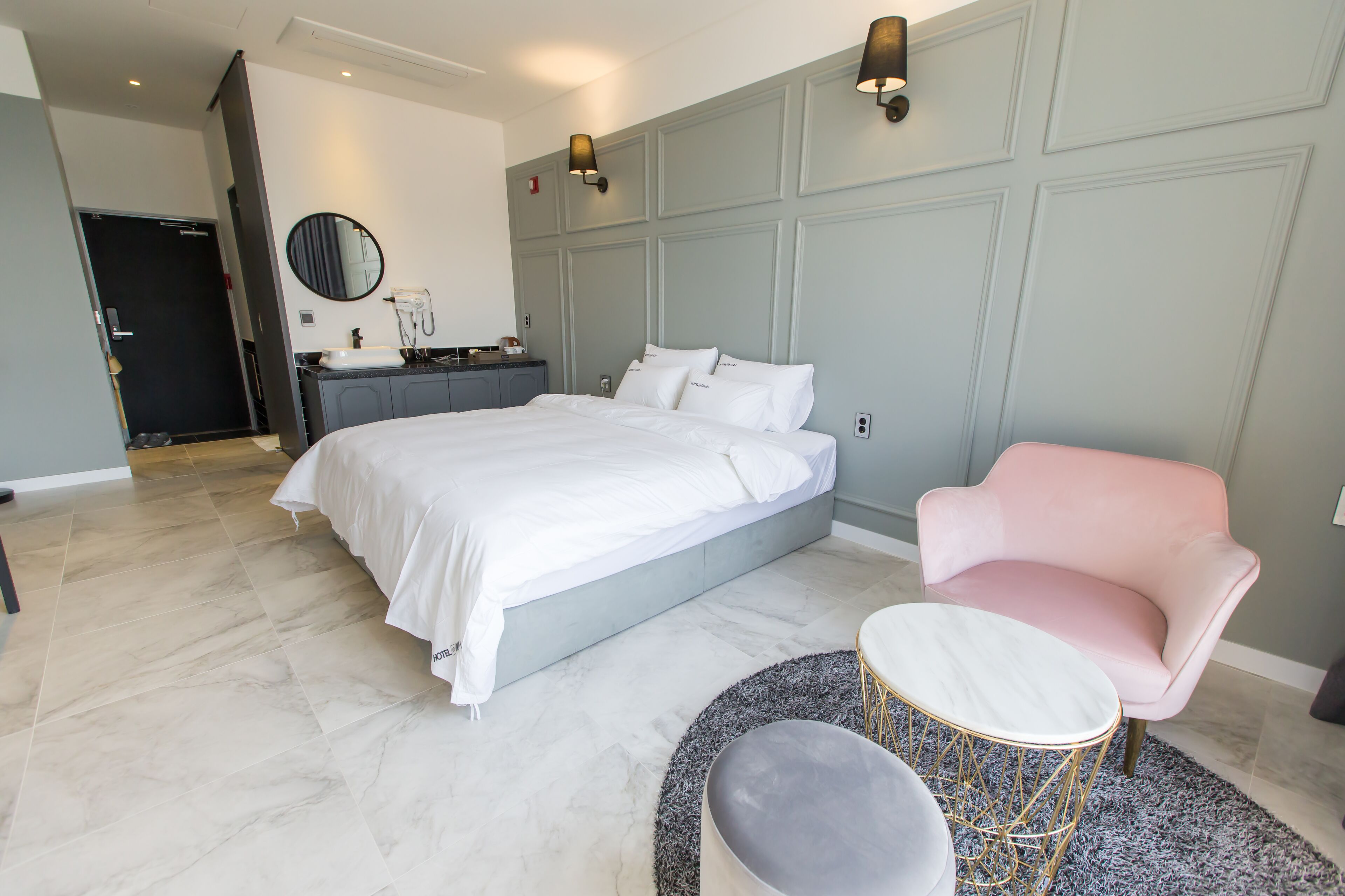 A modern bedroom with a large bed, a vanity with a mirror, a pink armchair, and a small round table.